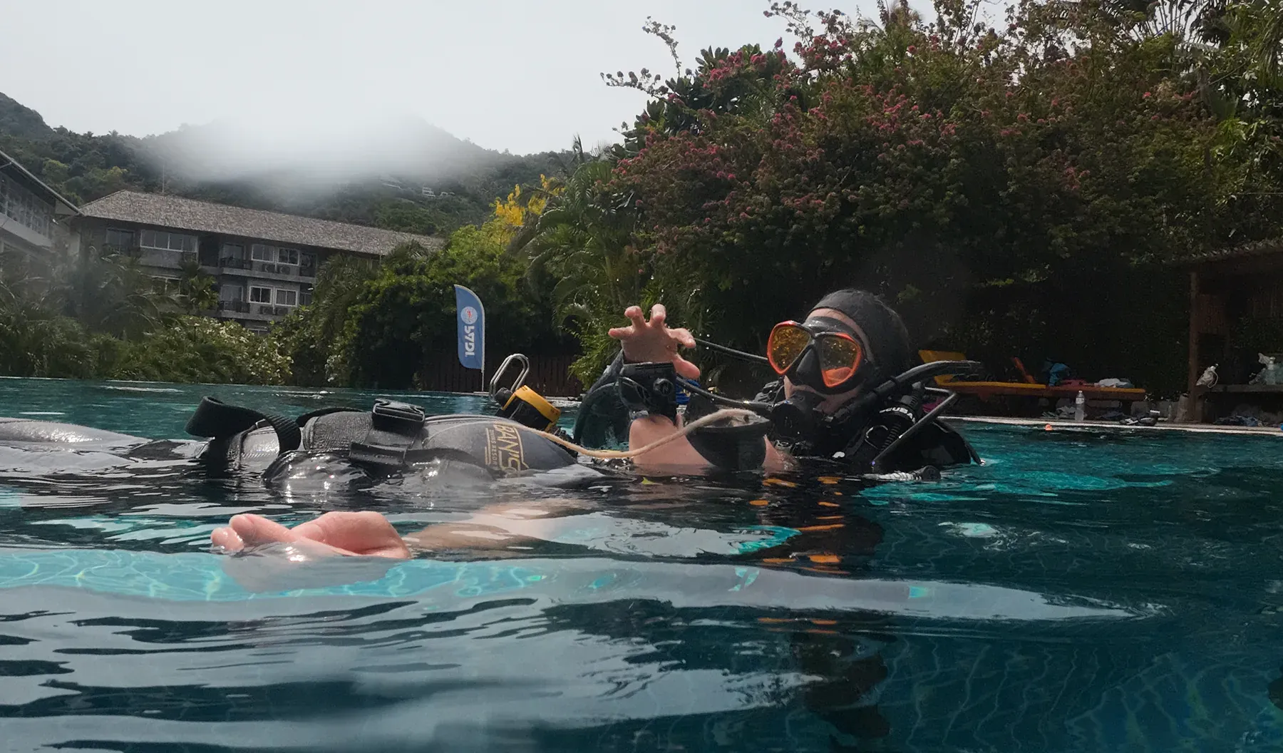 A divemaster in the training pool at Ban's Diving Resort on Koh Tao, running surface skills with a trainee, the hillside and resort grounds rising behind.