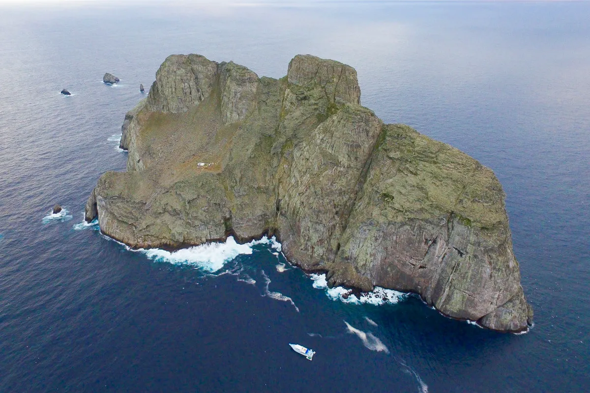 Aerial view of Malpelo Island in the Colombian Pacific, sheer basalt cliffs rising from deep blue water, a single liveaboard yacht moored at the southern point.