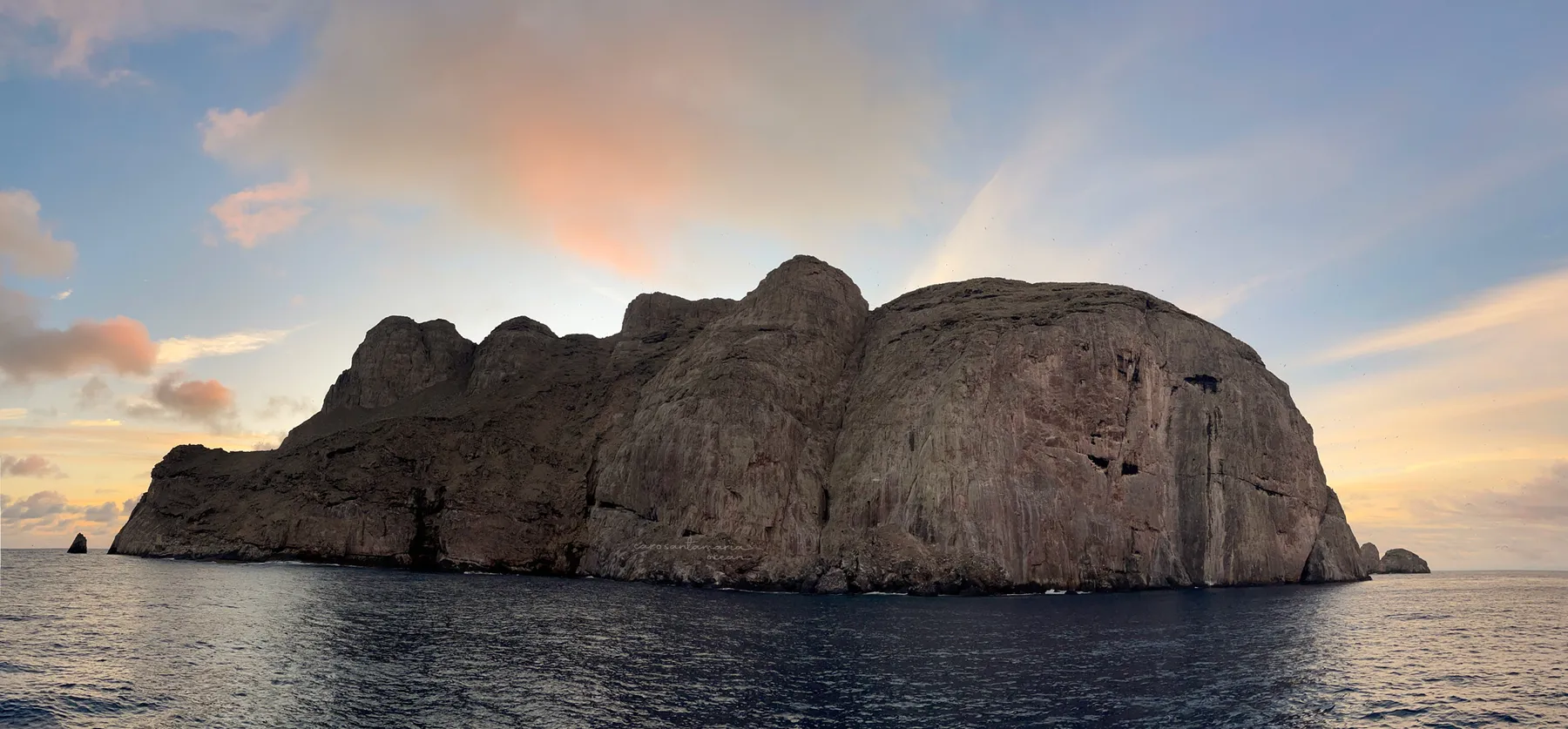 Wide panoramic view of Malpelo Island at dawn, sheer basalt cliffs rising from the Colombian Pacific under a pink and orange sky.