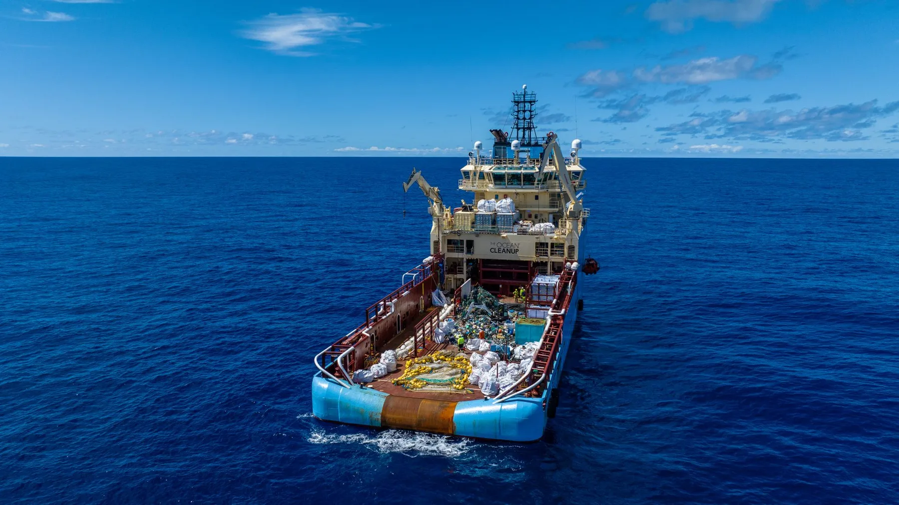 Vista aérea del buque de recolección de The Ocean Cleanup en medio del Pacífico, con la cubierta trasera cubierta de bolsas clasificadas de plástico y redes fantasma sacadas de la Gran Mancha de Basura del Pacífico, agua azul profunda abierta extendiéndose hasta el horizonte.