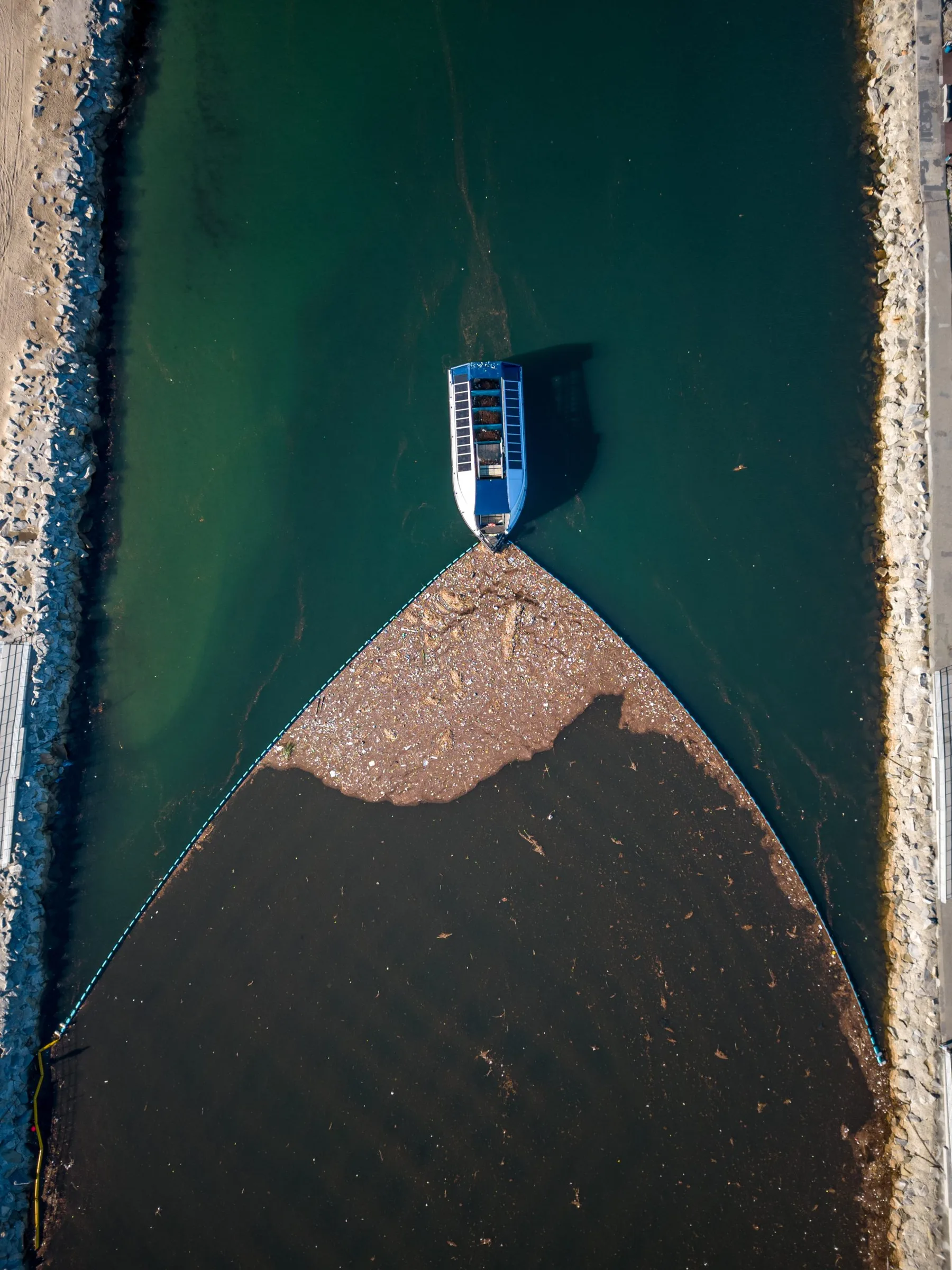 Vista aérea cenital de un Interceptor de Ocean Cleanup desplegado en la desembocadura de un río, la barrera flotante canalizando una densa balsa de plástico y desechos orgánicos hacia una embarcación de recolección alimentada por energía solar en el vértice.