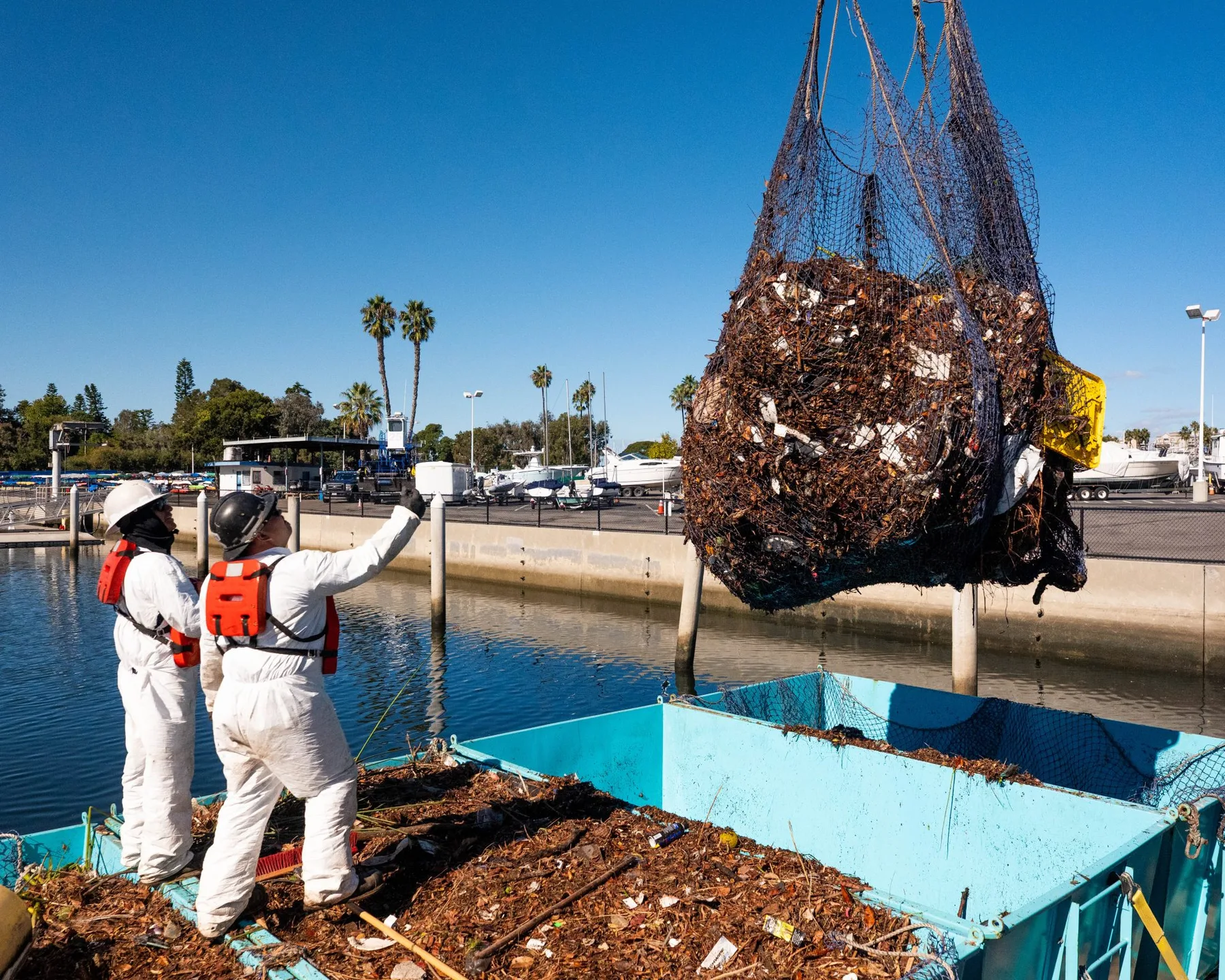 Dos trabajadores con trajes blancos de protección y chalecos salvavidas naranjas observan cómo una grúa levanta una gran red llena de plástico fluvial interceptado y desechos orgánicos sobre un muelle, junto a un contenedor de recolección de color turquesa.