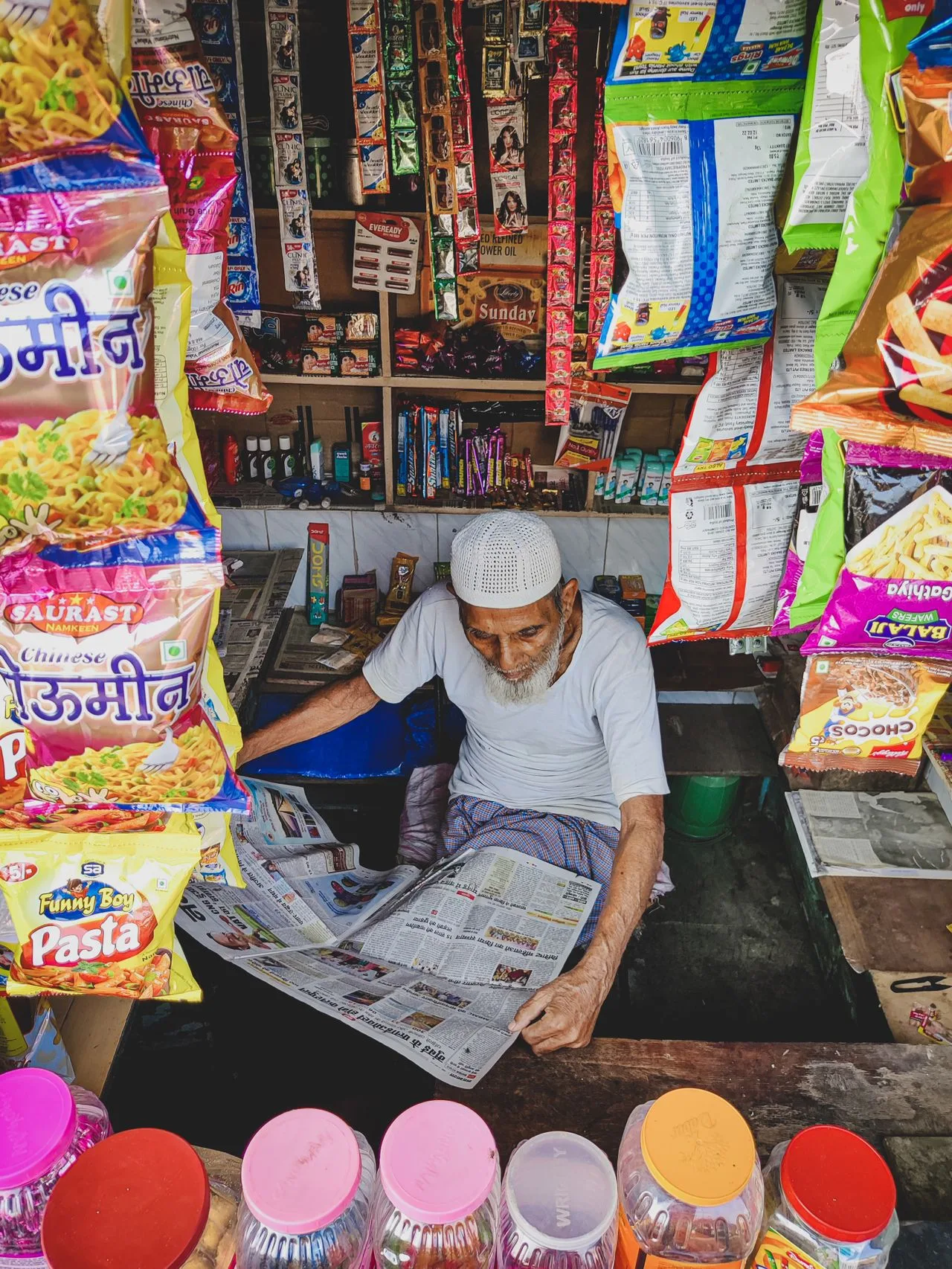 An elderly man reads a newspaper in a small snack stall surrounded by merchandise with plastic packaging