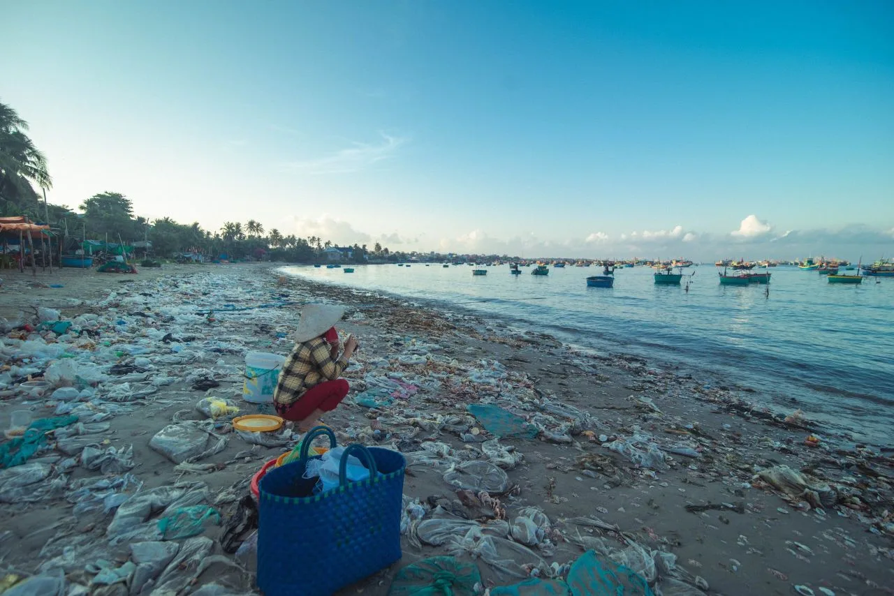 A beach covered with plastic pollution, highlighting environmental issues. Fishing boats are visible.