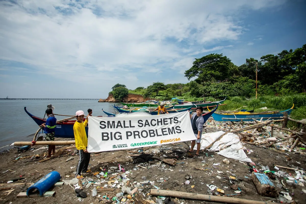 Residents of a coastal holding a banner that reads "small sachets. big problems" at a beach with small boats and surrounded by plastic pollution