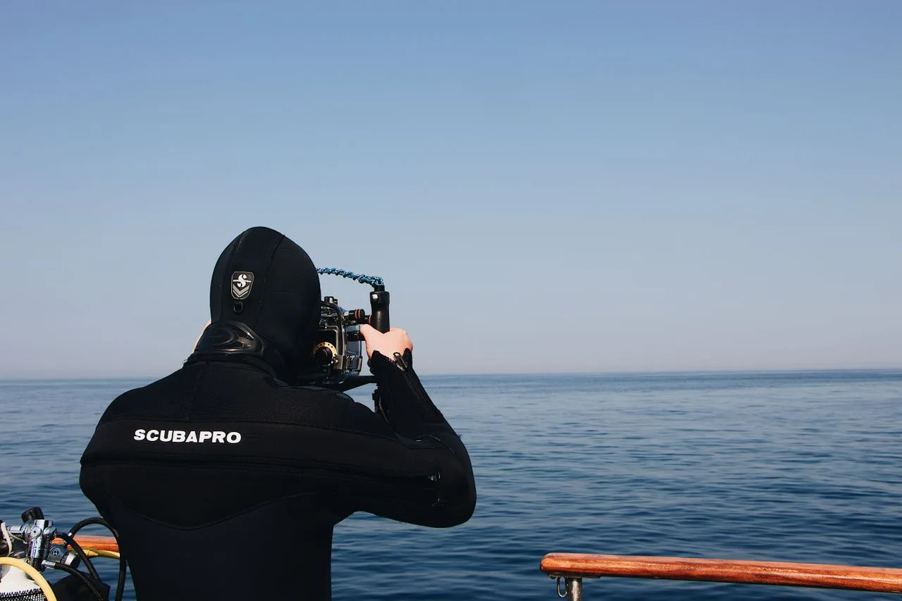 fotógrafo submarino con traje de neopreno y capucha Scubapro en un bote de buceo, sosteniendo una cámara